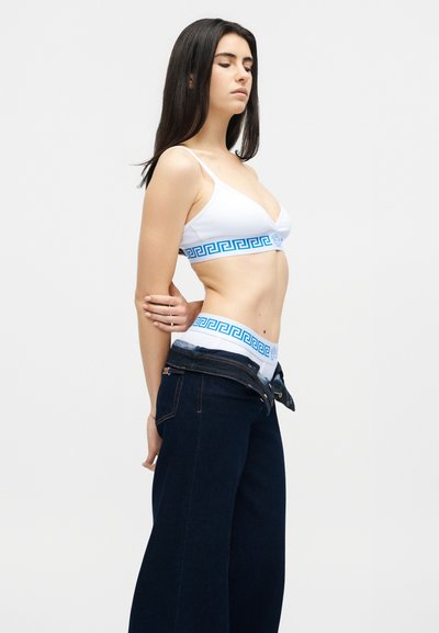 Γυναίκα που φοράει λευκό σουτιέν και εσώρουχα με μπλε γεωμετρικό περίγραμμα, τραβώντας προς τα κάτω σκούρα τζιν παντελόνια σε απλό ανοιχτό φόντο.