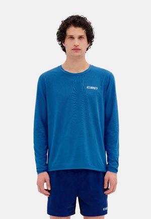 Man with curly hair wearing a blue long-sleeve shirt and matching shorts standing against a plain white background.