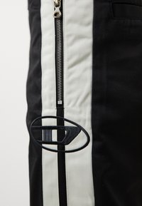 Gros plan sur un pantalon noir et blanc à rayures verticales, une fermeture éclair en métal, et un logo ovale noir brodé sur le côté gauche.