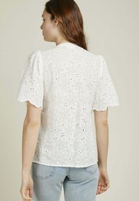 Blouse blanche en broderie anglaise avec des manches courtes bouffantes, des bords festonnés et des motifs floraux. Associée à un jean en denim bleu clair. Ajustement décontracté.