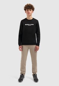 Zwarte sweatshirt met "BALLIN" in witte letters, beige broek en zwarte sneakers. De outfit heeft ribbelmanchetten en een relaxte pasvorm.
