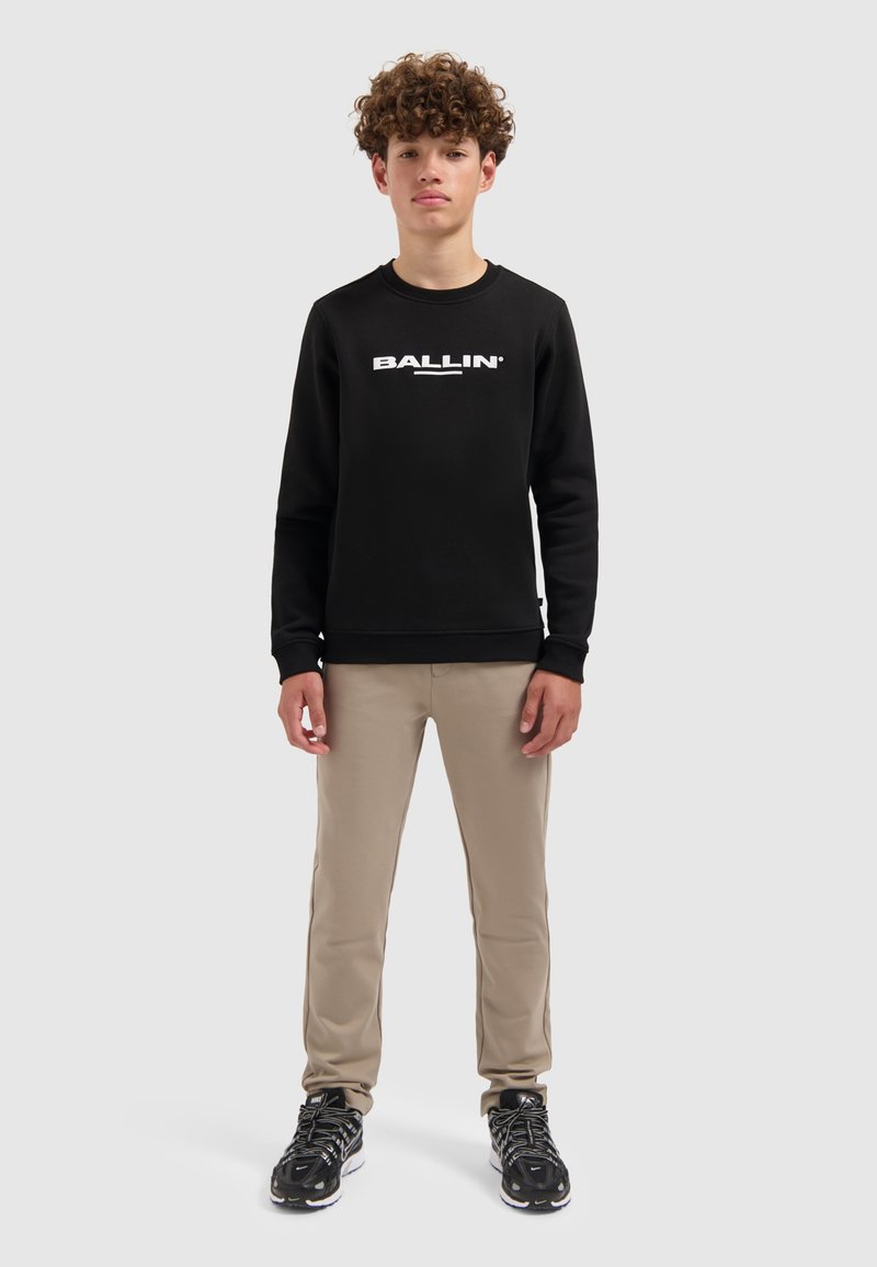 Zwarte sweatshirt met "BALLIN" in witte letters, beige broek en zwarte sneakers. De outfit heeft ribbelmanchetten en een relaxte pasvorm.