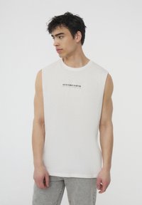 Camisetta bianca senza maniche con un collo a girocollo e una finitura testurizzata. Presenta testo stampato in nero, abbinata a pantaloni in denim grigio chiaro.