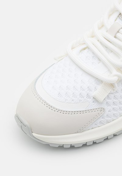 Lightweight sneaker featuring a white mesh upper, beige leather accents, textured surfaces, and a flexible rubber sole with traction patterns.