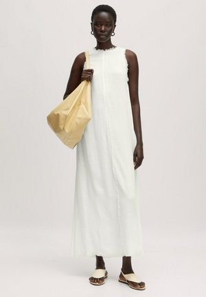 Vrouw draagt een mouwloze witte maxi-jurk, draagt een grote beige schoudertas en draagt beige sandalen, staand tegen een effen achtergrond.