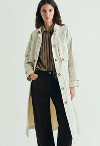 Kvinde iført en cremefarvet trenchcoat over en brunstribet skjorte og sorte bukser med vide ben, stående med den ene hånd i lommen.
