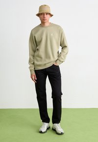 Lys oliven sweatshirt med et lille hvidt logo, sorte bukser, beige bøttehat og grå sneakers med sorte accenter, på et grønt gulv.