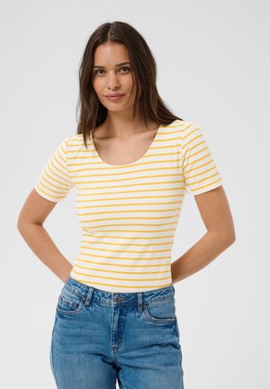 Jeune femme aux longs cheveux bruns portant un t-shirt blanc à manches courtes avec des rayures jaunes et un jean bleu, debout les mains dans le dos.