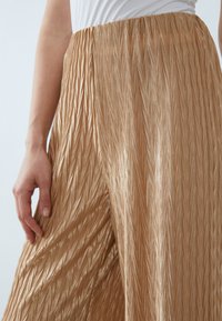 Pantalons plissés de couleur dorée fabriqués dans un tissu brillant et texturé avec une taille ajustée, présentant un motif en zigzag sur toute la surface.