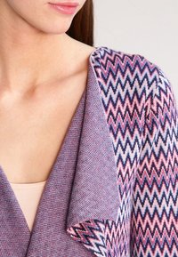 Teksturowany dzianinowy kardigan w odcieniach fioletu z motywem zygzaka w różowym i granatowym kolorze. Miękka tkanina z luźnym kołnierzem z przodu.