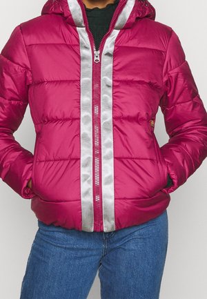 Personne portant une doudoune magenta avec une fermeture éclair argentée réfléchissante et un jean bleu, les mains dans les poches de la doudoune.