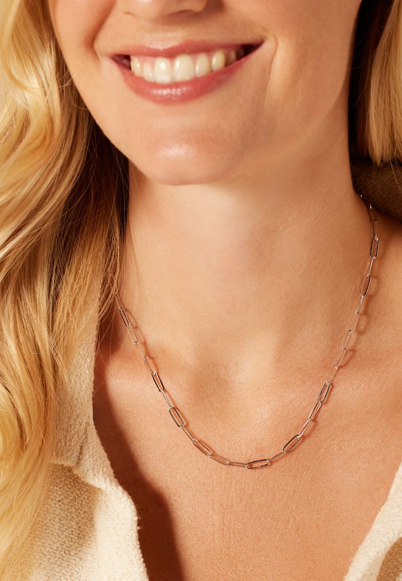 Zilverkleurige ketting met verlengde schakels in verschillende vormen en maten, liggend op een naakte nek met een gladde textuur.