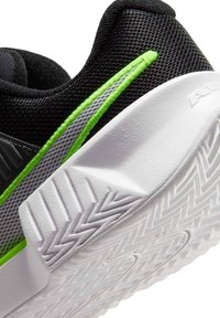 Μαύρο αθλητικό παπούτσι με ανώτερο μέρη από δίχτυ, πράσινες και γκρι ρίγες διακόσμησης, και λευκή, υφή σε λαστιχένια σόλα με μοτίβο ζιγκ-ζαγκ για καλύτερη πρόσφυση.