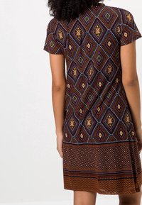 Femme portant une robe à manches courtes avec des motifs géométriques en losanges marron, violet et jaune, vue de dos sur fond blanc.