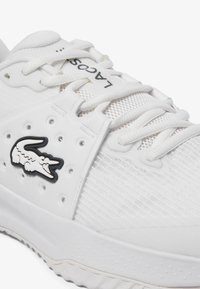 Scarpa sportiva bianca realizzata in materiali mesh e gomma, con lacci piatti, perforazioni e un logo di coccodrillo nero di lato.