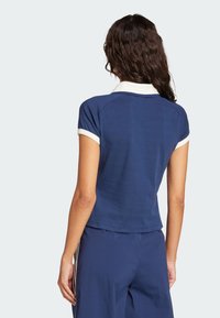 Polo majica tamnoplave boje s bijelim ovratnikom i naglascima na rukavima. Kratki rukavi, prilagođen dizajn i mekana tekstura. Prikazan stražnji pogled.