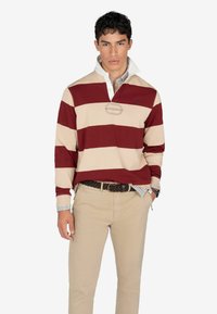 Polo a righe bordeaux e beige con colletto bianco, dotato di un logo cucito e un motivo a quadri sottostante, abbinato a pantaloni beige.
