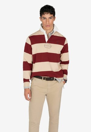 Camisa de polo a rayas color burdeos y beige con cuello blanco, que presenta un logo bordado y un diseño a cuadros debajo, combinada con pantalones beige.