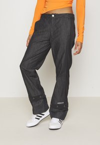 Svart texturerad tygbyxor med lös passform, tryckknappar vid anklarna och Adidas-logotypen visad nära fållen.