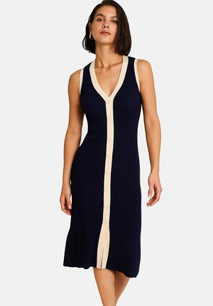 Femme portant une robe midi bleu marine sans manches à boutons avec une bordure beige le long du col, des boutons et des côtés, debout sur fond blanc.