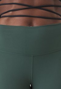 Green high-waisted leggings with a smooth texture and double stitching. Black crisscross straps accentuate the upper part.