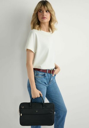 Blondynka w białej krótkiej bluzce i niebieskich dżinsach trzyma czarną teczkę na dwa uchwyty, stojąc na tle jednolitego tła.