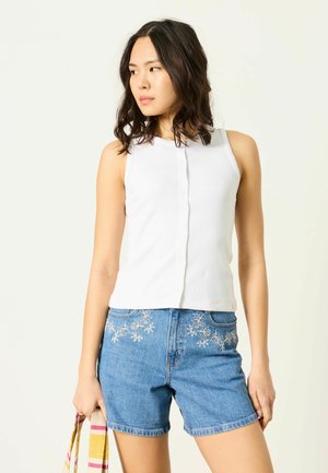 Žena v bielom bezrukávovom topu a modrých rifľových šortkách s kvetinovou výšivkou, drží pruhovaný uterák a pozerá sa doľava.