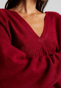 Femme portant un pull en maille rouge foncé à col en V avec des manches bouffantes et un délicat collier en argent orné d'un pendentif barre horizontale.