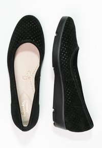 Zapatos de ante negro de tipo slip-on con parte superior perforada, puntera redonda, suela baja negra y plantilla beige con un diseño de lazo cosido.
