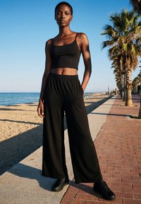 Young woman wearing black crop top and wide-leg pants standing on a sunny beach promenade with palm trees and sea in the background.