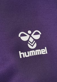 Wit gestileerd hommel-logo en de tekst "hummel" gedrukt op paarse stof, die het sportkledingmerk Hummel vertegenwoordigen.