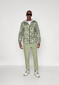 Žalios švieskamufliažinės striukės su priekiniu užtrauktuku ir gobtuvu derinys su šviesiai žaliomis kelnėmis ir neutralaus stiliaus sportbačiais. Modelis dėvi tamsius akinius nuo saulės.