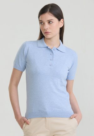 Kvinde iført en lyseblå strikket polo med korte ærmer og knapper, stående med hænderne i lommerne på beige bukser mod en ensfarvet baggrund.