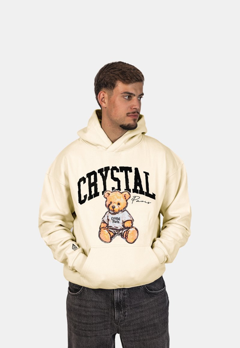Felpa crema con un orso cartone animato e la scritta "Crystal Paris". Tessuto morbido, vestibilità over, e tasca a marsupio, indossata con jeans scuri.