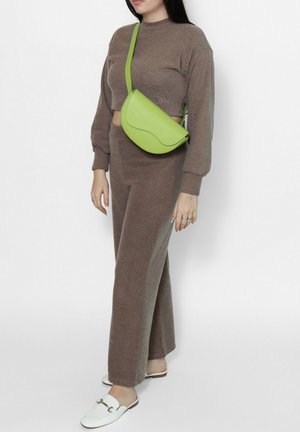 Femme portant un haut en maille marron et un pantalon assorti, avec des chaussures blanches faciles à enfiler et un sac bandoulière vert vif, debout devant un fond clair.