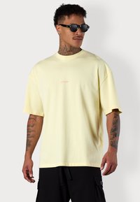 Helles gelbes übergroßes T-Shirt mit Rundhalsausschnitt und kurzen Ärmeln. "ESTEEM"-Logo in Pink auf der Vorderseite. Glatter Baumwollstoff.