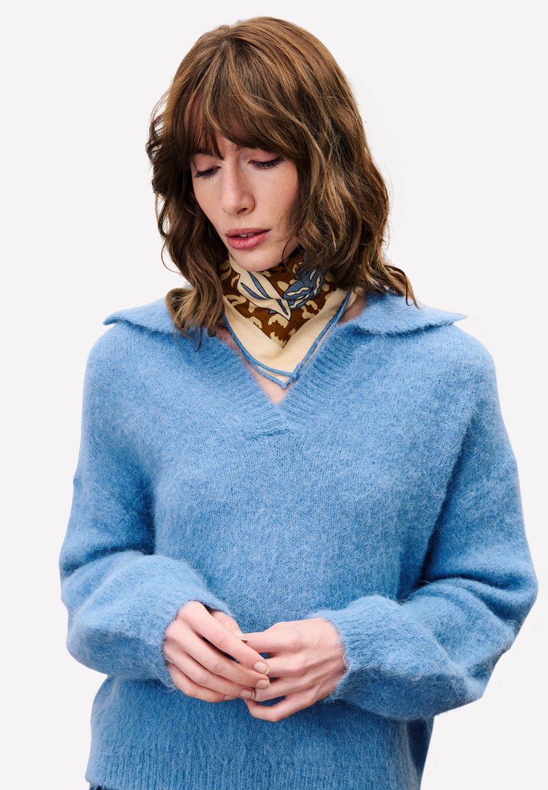 Hellblaues Sweater aus weichem, flauschigem Material, mit V-Ausschnitt, weiten Ärmeln und einem Kragen. Ein gemusterter brauner Schal ist um den Hals gebunden.