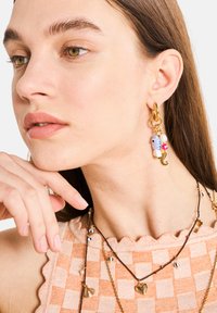 Des boucles d'oreilles en or en forme de cerceau arborent un charme en forme de poisson bleu, une perle blanche et un accent rose. Le modèle porte un haut à motif à carreaux pêche avec des bords festonnés.