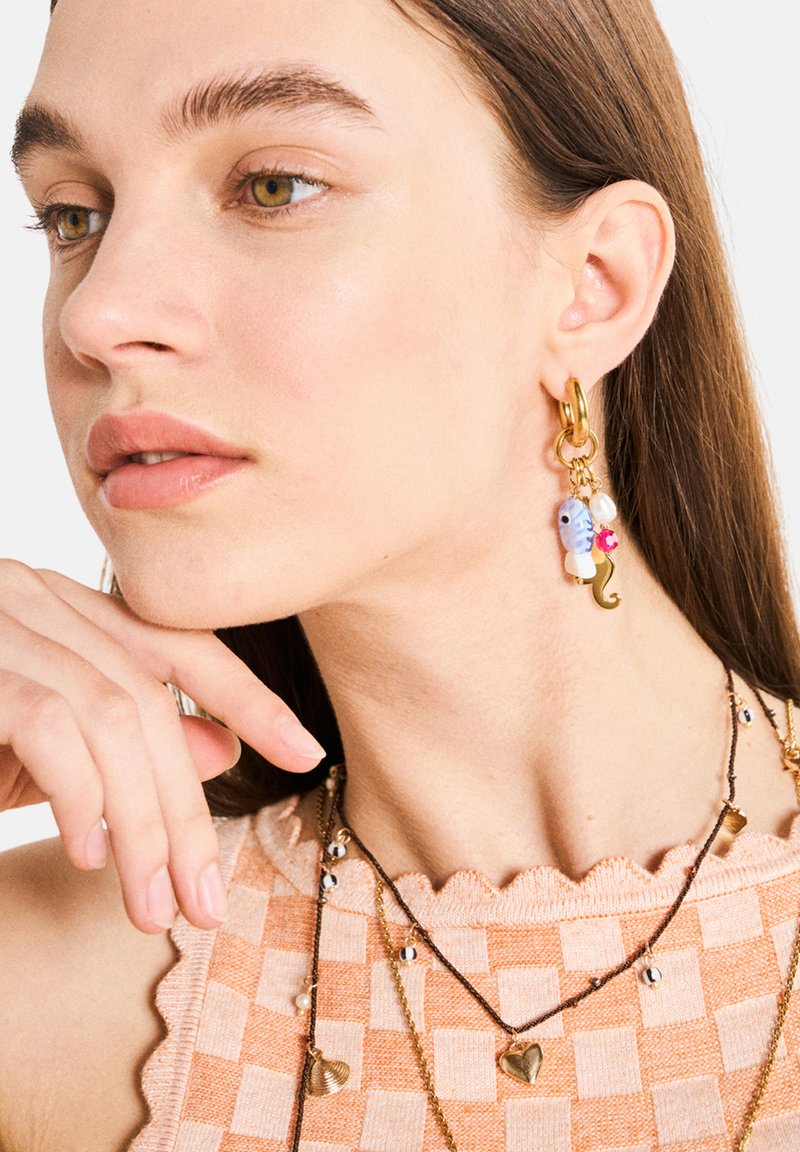 Des boucles d'oreilles en or en forme de cerceau arborent un charme en forme de poisson bleu, une perle blanche et un accent rose. Le modèle porte un haut à motif à carreaux pêche avec des bords festonnés.