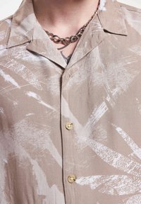 Beige Kurzarmhemd mit weißem abstraktem Pinselstrichmuster, ausgestattet mit einem spitzen Kragen und zwei goldenen Knöpfen auf der Vorderseite.