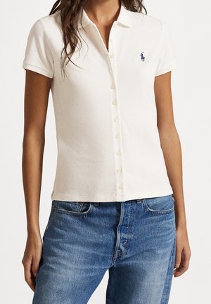 Kvinde iført en tætsiddende hvid poloshirt med knapper og korte ærmer samt blåt broderet logo, kombineret med blå denimjeans.