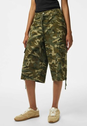 Parte inferior del cuerpo de una persona con pantalones cortos cargo de camuflaje y zapatillas beige con cordones blancos sobre un fondo claro.