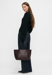 Ciemnobrązowa torba na ramię o gładkiej teksturze, z delikatnymi wytłoczonymi wzorami i dwiema długimi rączkami, trzymana obok osoby w czarnym swetrze.