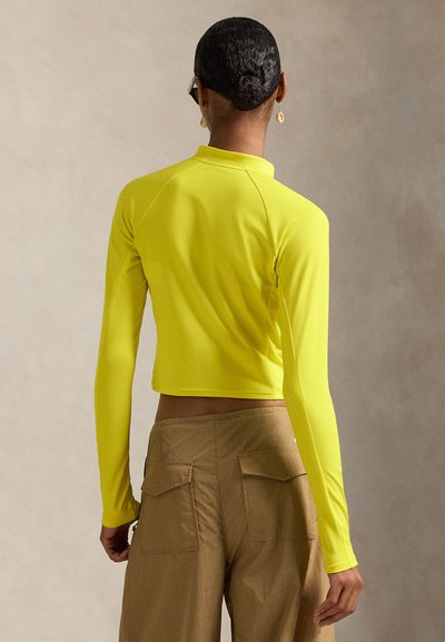 Φωτεινό κίτρινο κοντό τοπ με μακριά μανίκια και ψηλό γιακά, από ελαστικό υλικό, συνδυασμένο με καφέ παντελόνι τύπου cargo με διπλές πίσω τσέπες.