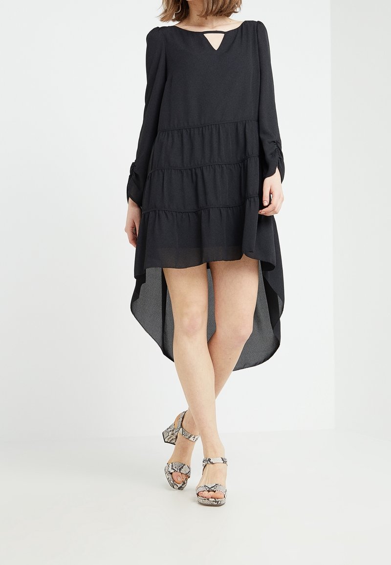 Femme portant une robe noire à manches longues, coupe asymétrique avec jupe à volants et sandales à talons hauts à motif peau de serpent, debout sur fond blanc.