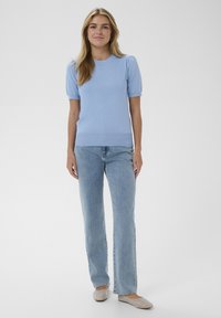 Femme debout portant un haut à manches courtes bleu clair, un jean délavé clair et des ballerines beiges ornées de perles, sur un fond blanc uni.