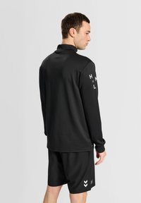 Schwarzes langärmliges Sporthemd mit hohem Kragen und weißem Branding auf dem Ärmel, kombiniert mit schwarzen Shorts mit einem weißen Chevron-Muster.