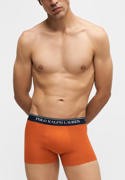 Άνδρας χωρίς πουκάμισο που φορά πορτοκαλί boxer Polo Ralph Lauren, στέκεται με τα χέρια πίσω από την πλάτη σε ένα απλό φωτεινό φόντο.