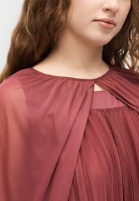 Νεαρό άτομο με μακριά σγουρά μαλλιά φοράει πλισέ, διαφανές φόρεμα σε ροζ απόχρωση με στρογγυλή λαιμόκοψη και μικρή λεπτομέρεια με άνοιγμα.