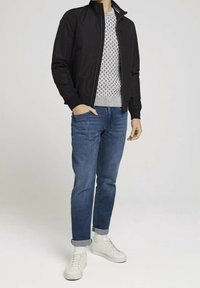 Černá lehká bunda, šedý texturovaný svetr, modré džíny a bílé tenisky. Oblečení má přední zip a manžety na kalhotách.